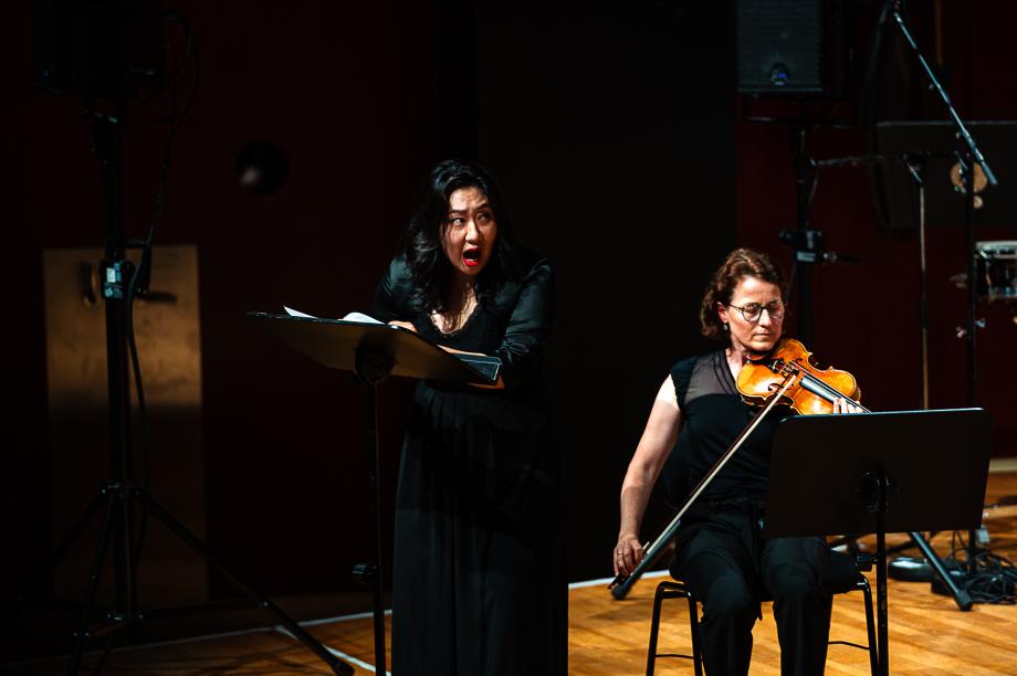Szenisch agierende Sängerin, in schwarz gekleidet, die ihren Blick seitlich auf die Bühnenmitte fokussiert.. Im Hintergrund ist eine Violinistin zu sehen.