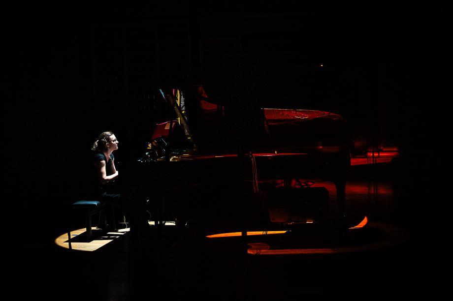Karolin Schmitt-Weidmann konzertierend am Flügel. Die Beleuchtung ist auf einen schmalen Kegel auf die Noten ausgerichtet. Die Dunkelheit des Konzertsaales und die Konturen des Flügels sind kaum zu unterscheiden.