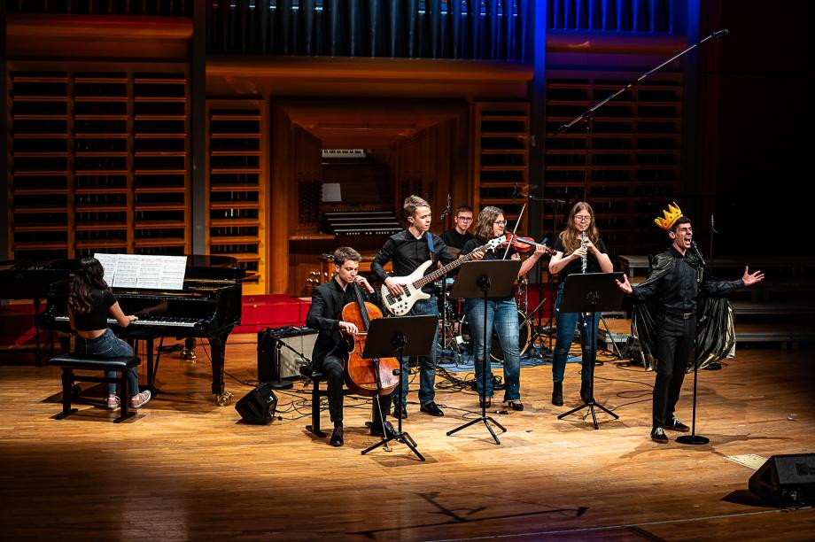 Schulmusikkonzert 2024: Studentin am Flügel, eine Band und ein Student mit einer Krone auf dem Kopf, der singt.