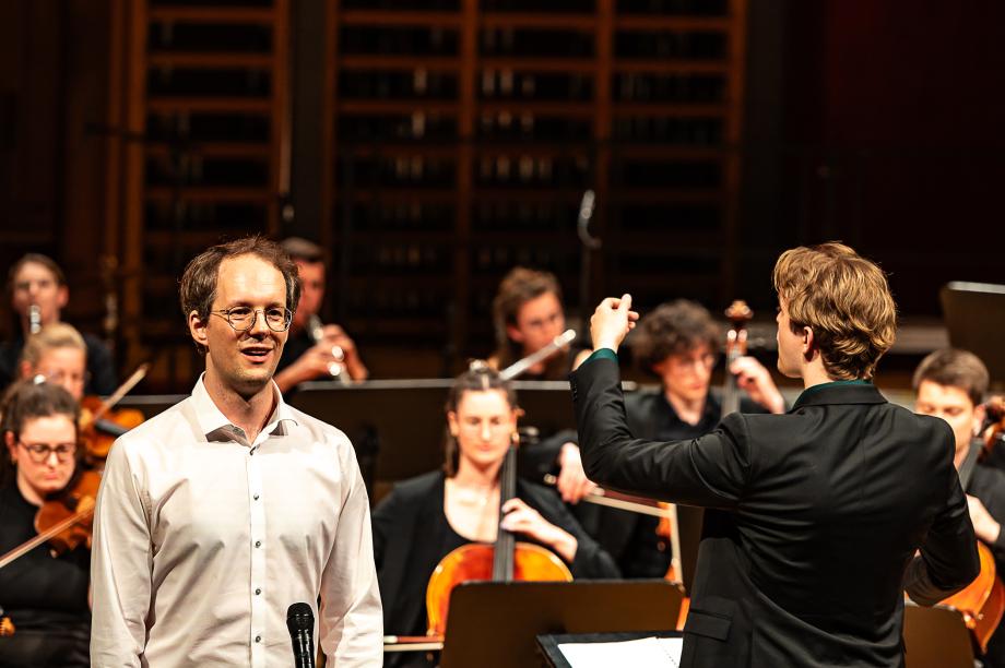 Schulmusikkonzert 2024: Im Hintergrund Studierende im Orchester. Im Vordergrund: ein Student der singt und einer, der dirigiert.