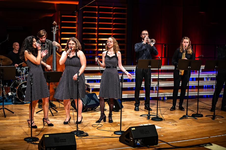 Schulmusikkonzert 2023: Band auf Bühne. Drei Studentinnen in schwarz-weiß gepunkteten Kleidern singen. 