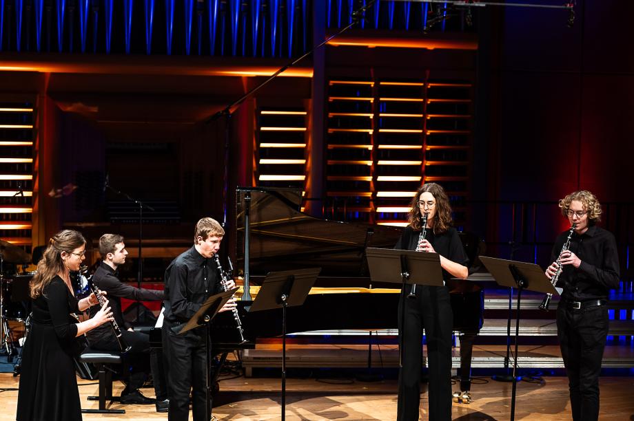 Schulmusikkonzert 2023: Im Hintergrund ein Student am Flügel. Im Vordergrund: vier Studierende an Notenständern spielen Klarinette.