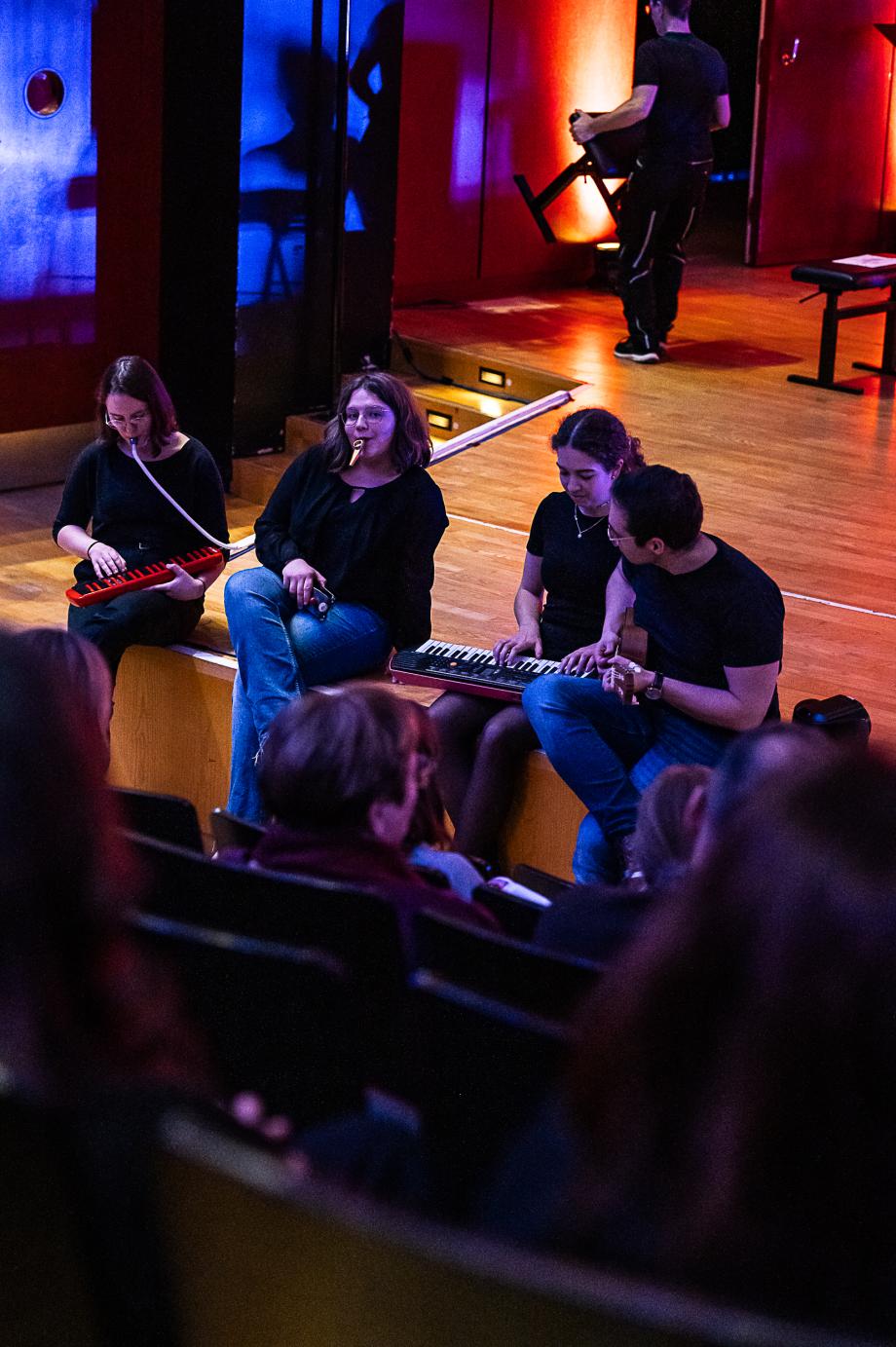 Schulmusikkonzert 2023: Studierende sitzen auf Bühnenrand zum Publikum hin gewendet und musizieren.
