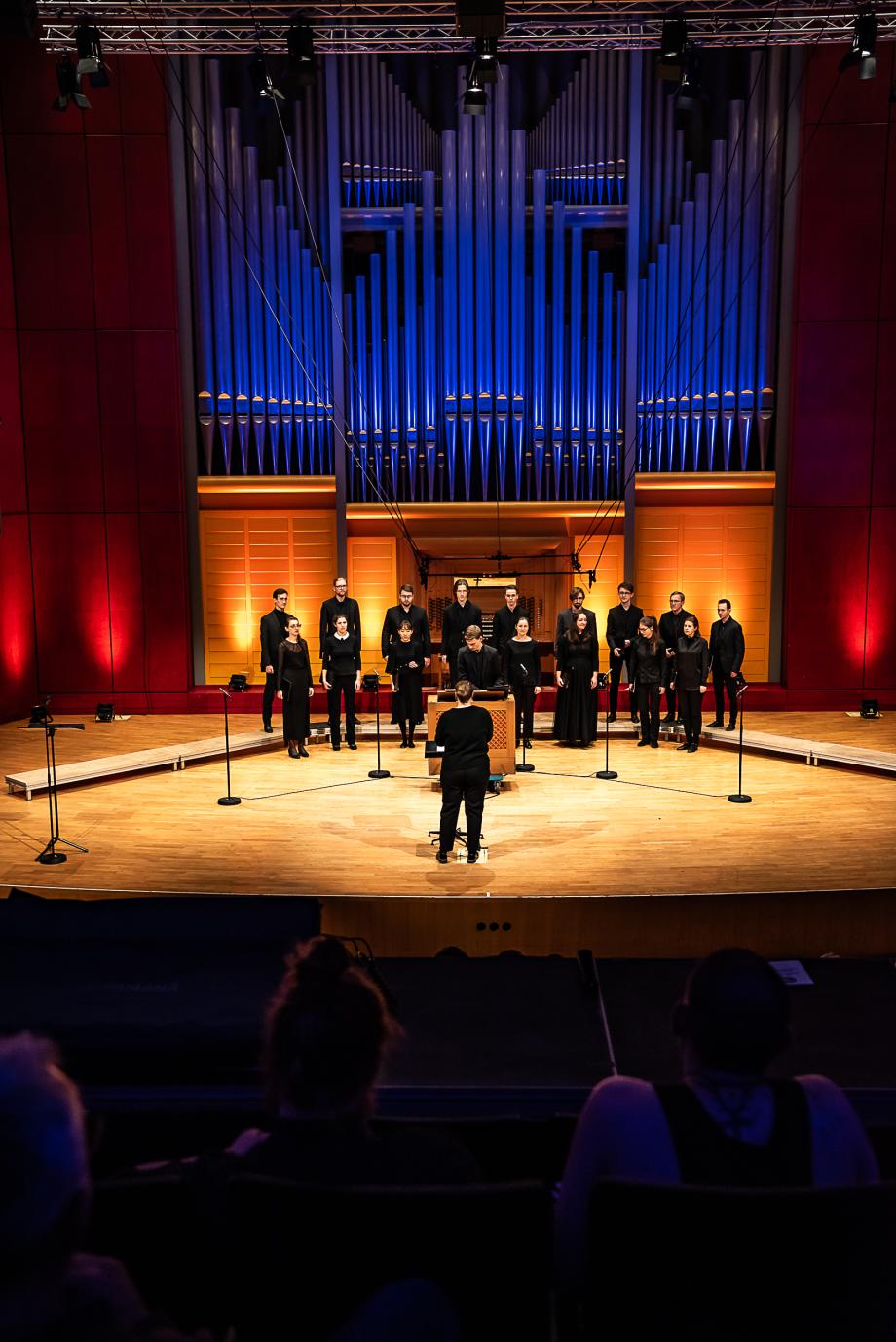 Das sechzehnköpfige Vokalensemble vor dem Orgelprospekt der Konzertsaalorgel. Mit dem Rücken zum Publikum steht die Dirigentin. Umgeben vom Vokalensembles ist der Studierende an der Truhenorgel.