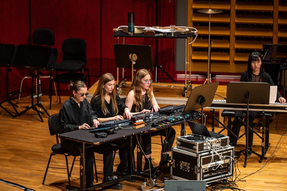 Dret Studierenden sitzen an einem Tisch mit unterschiedlichen Pulten. An einem gesondert Tisch sitzt eine Studierende mit Keyboard. Zwischen den Tischen sind Dutzende von Kabeln verlegt.