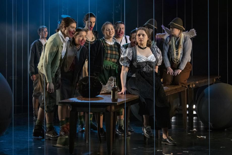 Studierende der Opernschule in einer Bühnenszene. Eine Darstellerin in Dirndl sitzt auf einem Tisch in der Mitte und die Gruppe von Studierenden lacht sie spöttisch von hinten an.