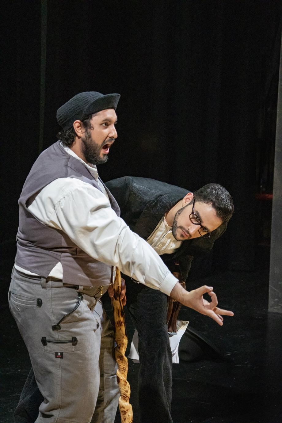 Zwei Sänger der Opernschule in einer Bühnenszene. Der eine singt im Vordergrund und formt mit Daumen und Mittelfinger einen Kreis, den der andere Sänger verwundert betrachtet.