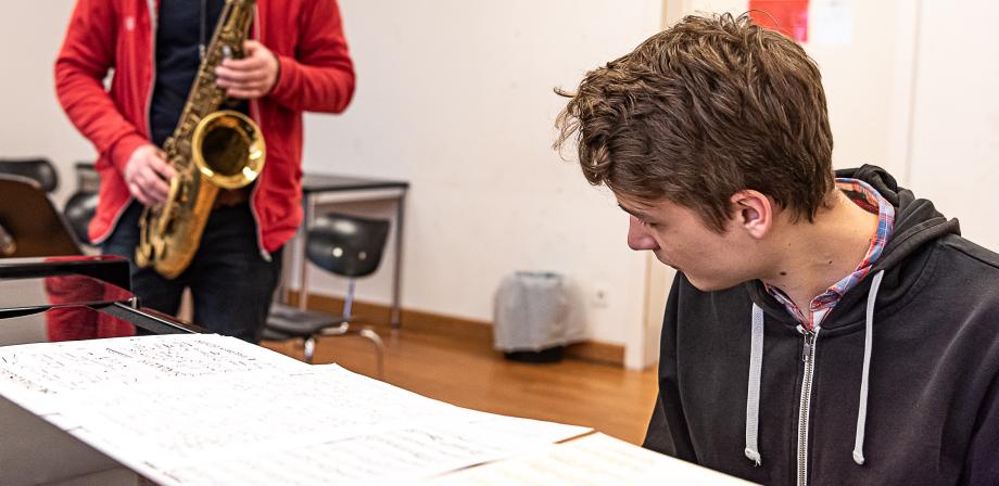 Student sitzt am Klavier. Sein Blick ist gesenkt, im Hintergrund spielt jemand Saxophon.