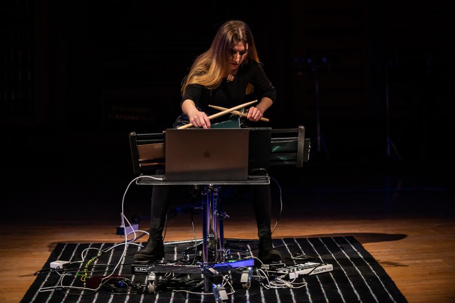 Schlagzeugerin mit Holzschlägeln, einem ausgezogenen Notenpult und Laptop. Auff dem Boden um sie herum sind viele Effektgeräte angeschlossen.