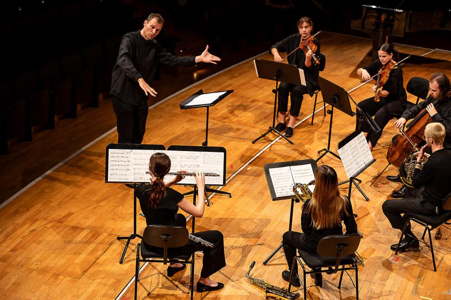 Studierende an Streich-und Blasinstrumenten auf Bühne, dirigiert von Christof M Löser.