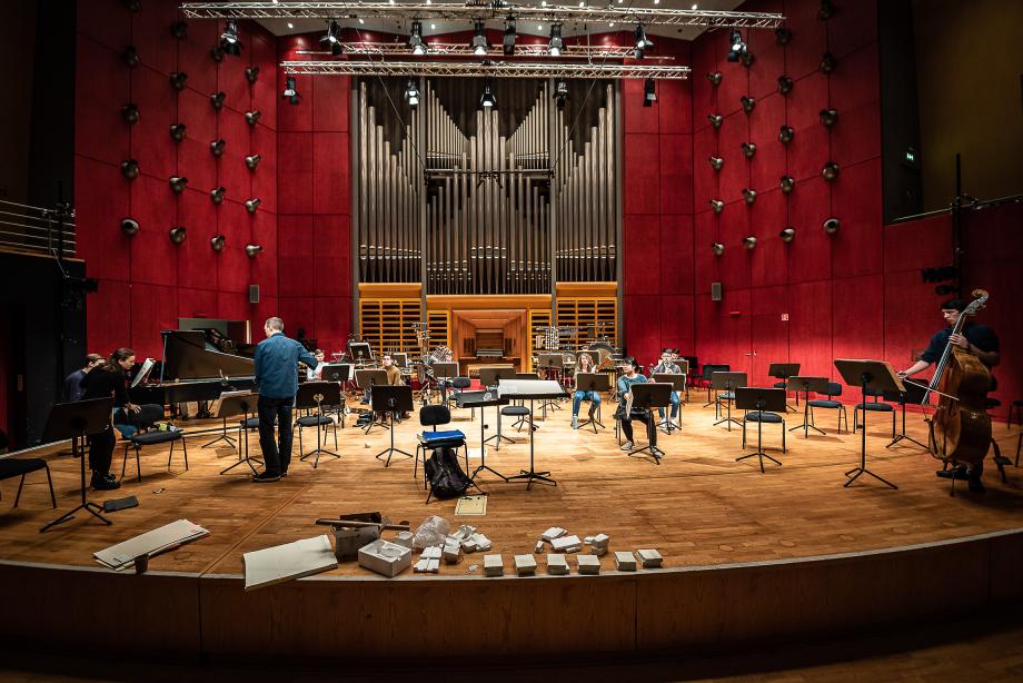 Blick vom Zuschauerraum auf die Bühne des Werkstattfestivals. Christ M Löser ist im Gespräch. An der Bühnenkante liegen viel kleinere, weiße Styroporstapel. Das Podium ist mit einem Orchesteraubfau für ein Kammerensemble versehen.
