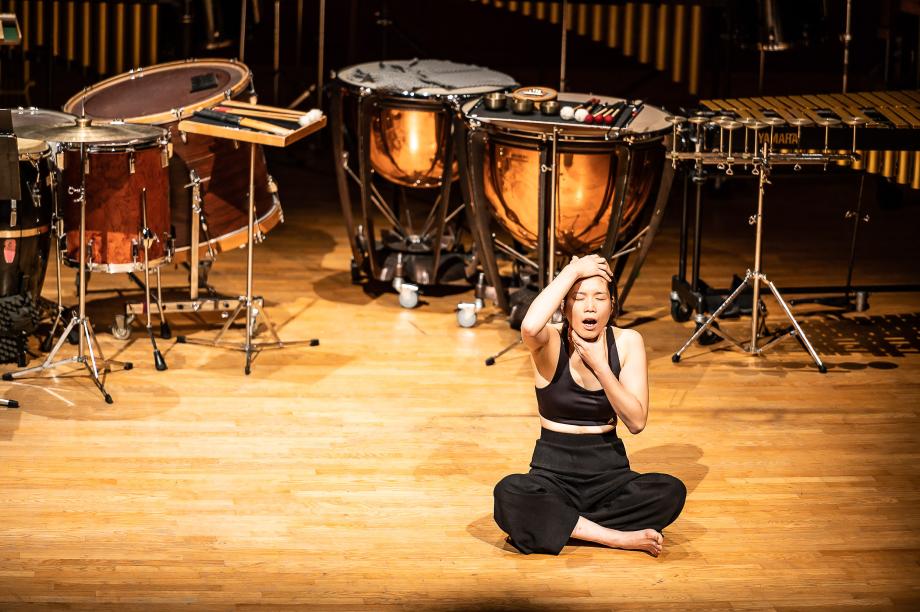 Szenisch agierende Schlagzeugstudentin, auf dem Podium sitzend, vor einem umfangreichen Schlagzeugaufbau.