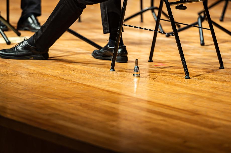 Detailaufnahme eines Blechbläsers, wo nur die Füße, der Orchesterstuhl und ein silbernes Mundstück zu sehen ist.