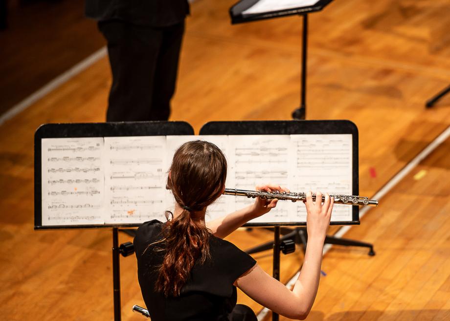 Musizierende Querflötistin, von hinten fotografiert. Sie spielt aus zwei nebeneinanderstehenden Notenpulten, um ihre umfassende Einzelstimme umsetzen zu können.