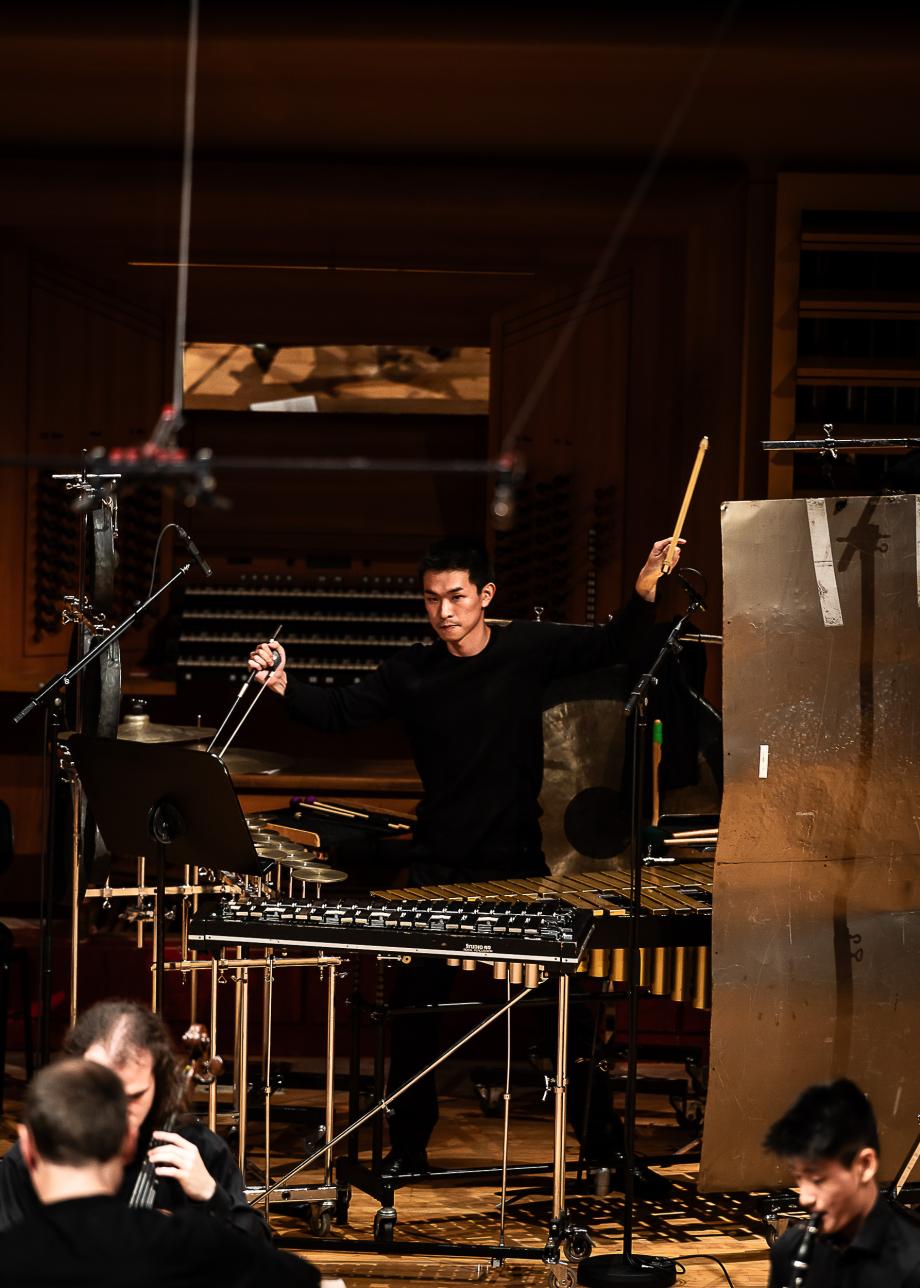 Agierende Schlagzeugstudent, der mit der rechten Hand und einem Kontrabassbogen Gongs streicht und mit einem Holzschlägel der linken Hand ein Wellblech schlägt. Er ist umgeben von musizierenden Studierenden.