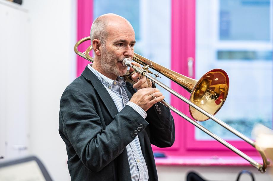 Portraitaufnahme des Posaunenprofessor Henning Wiegräbe im Unterrichtszimmer. Im Hintergrund dominiert farblich der pinke Fensterrahmen.