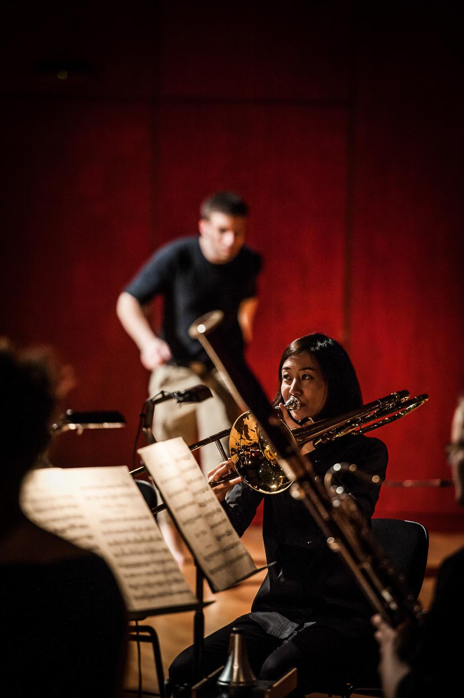 Detailaufnahme einer Posaunistin. Verschwommen ist eine Fagottistin zu erkennen und im Hintergrund ein Schlagzeuger.