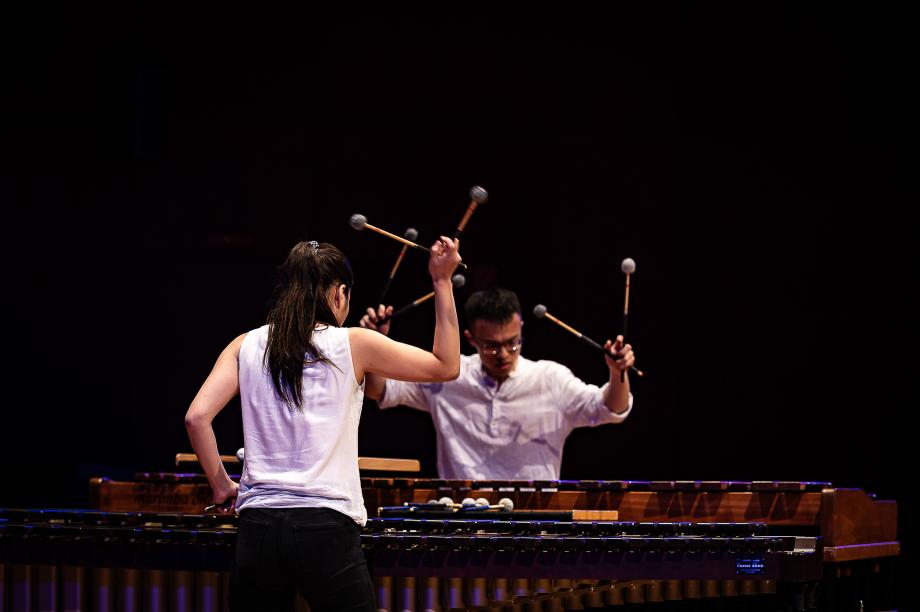 Schkagzeugstudierende an Marimbaphinen. Die stehen sich genüber, haben ihre arme erhoben und die Scchlägel zu einem V gebildet.