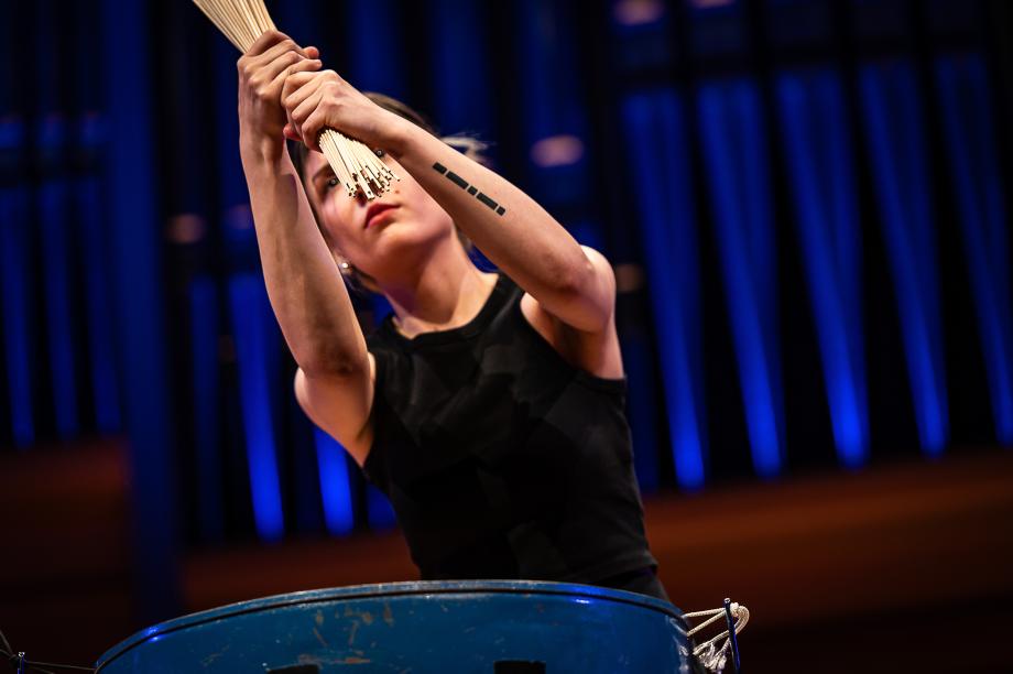 Schlagzwugstudentin an einer großen Trommel. Sie umfasst mit ihren beiden Händen ein Bündel Mikadostäbe. Auf dem linken Unterarm ist eine größere Tätowierung mit unterschiedlich langen Balken zu sehen.