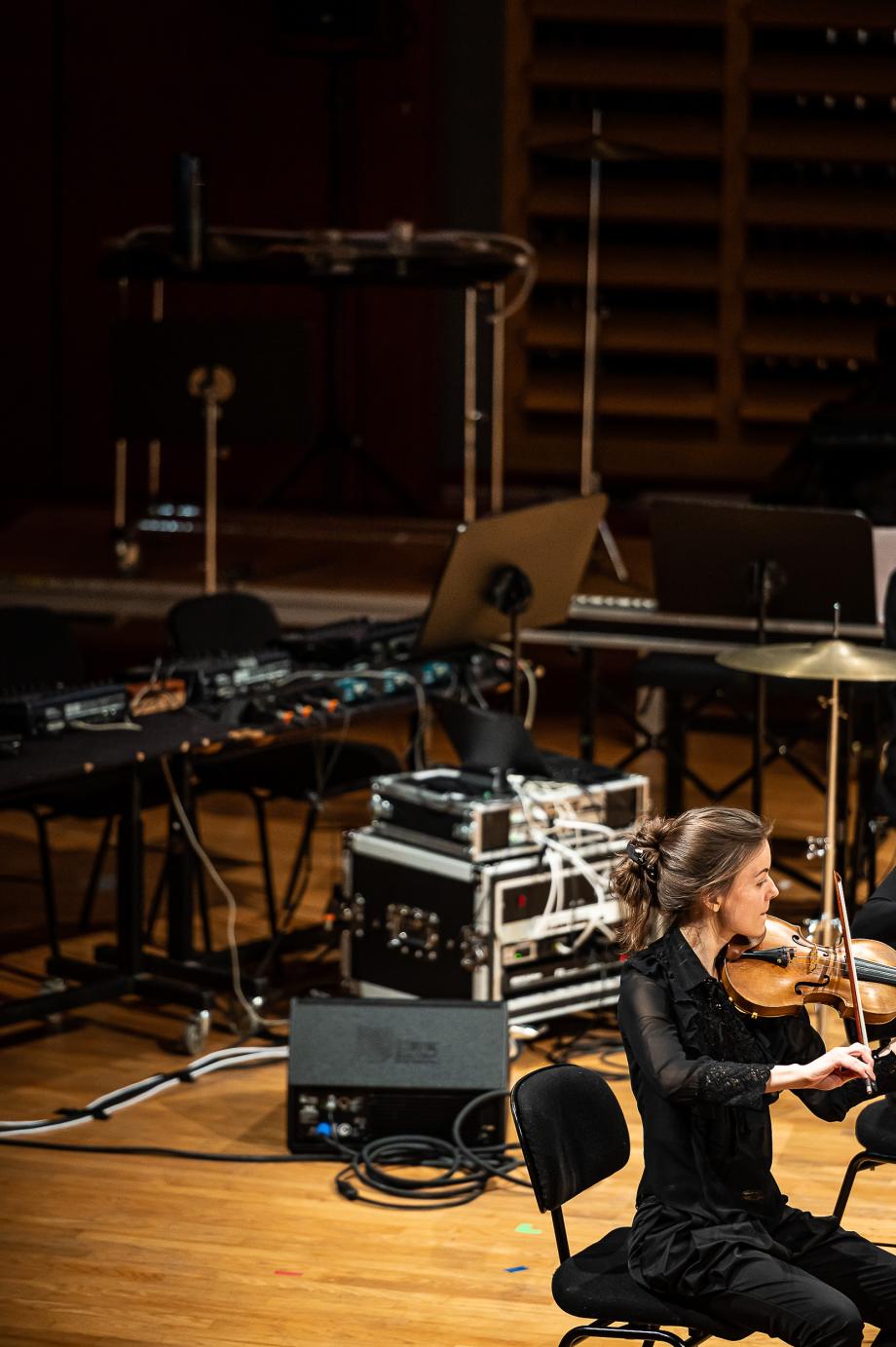 Geigenstudentin beim Werkstattfestival im Konzertsaal. Im Hintergrund sind verschiedene Verstärker und eine PA-Anlage auf der Bühne installiert.