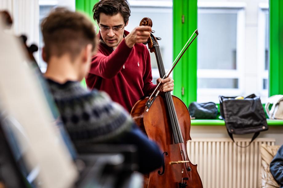 Celloprofessor Tristan Cornut beim Unterrichten. Mit ausgestreckten Arm und Zeigefinger deutet er den Zielpunkt der Phrase an. Der musizierende Studierende ist von hinten zu sehen.