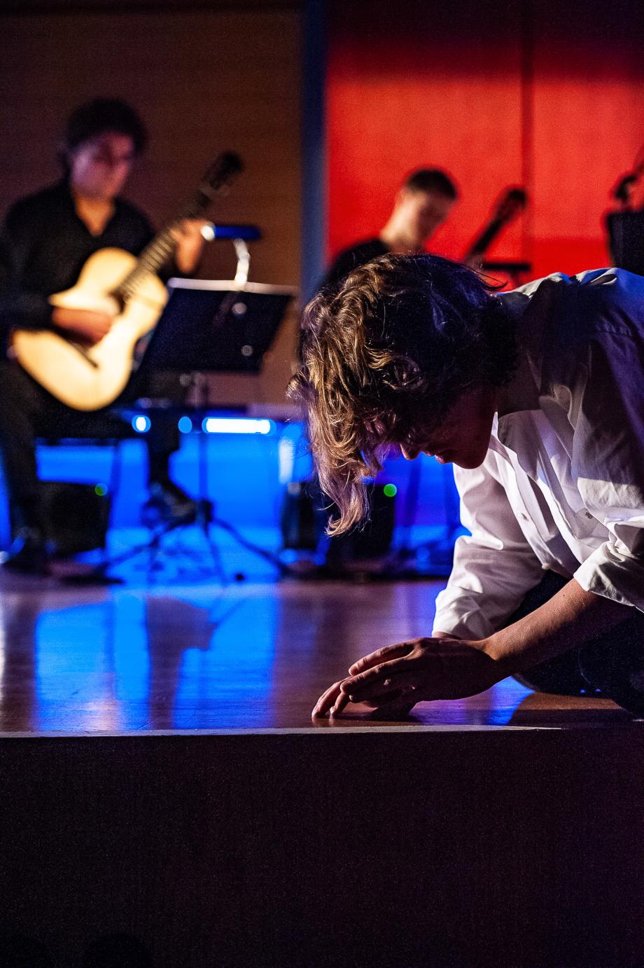 SChauspieler kniend im Vordergrund. Im Hintergrund ist ein Gitarrenensemble zu erkennen.