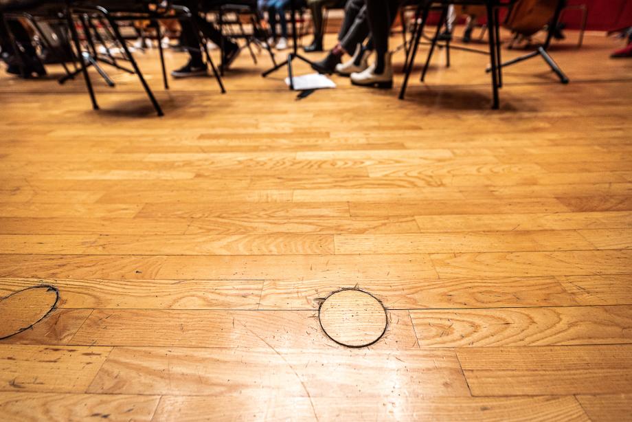 Detailblick auf die helle parkettvorbühne. Im Hintergrund sind Notenständer, Stuhlbeine und Füße von musizierenden Studierenden zu sehen.