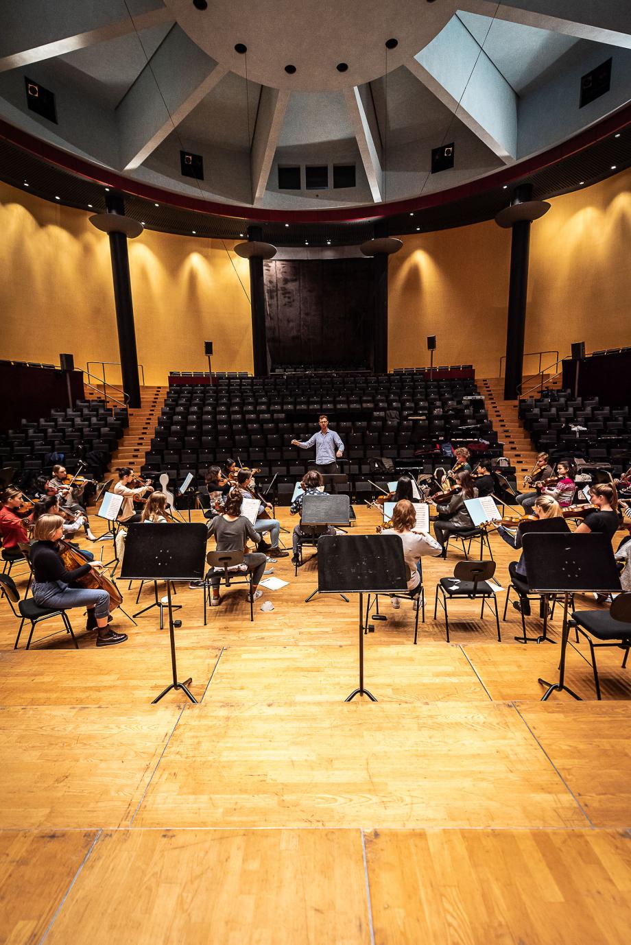 Blick von der Orgel auf ein musizierenden Ensemble unter der Leitung von Christof M Löser. Im Hintergrund ist der leere Zuschauerraum sichtbar.