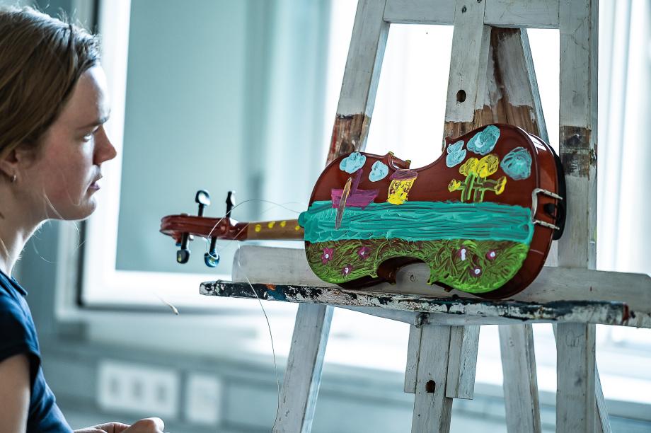 Studentin blick auf eine mit Acrylfarbe bemalte Violine. Zu erkennen ist ein Fluss, ein SChiff und eine Blumenwiese. Die Violine ist auf einer weißen Staffel liegend positioniert.