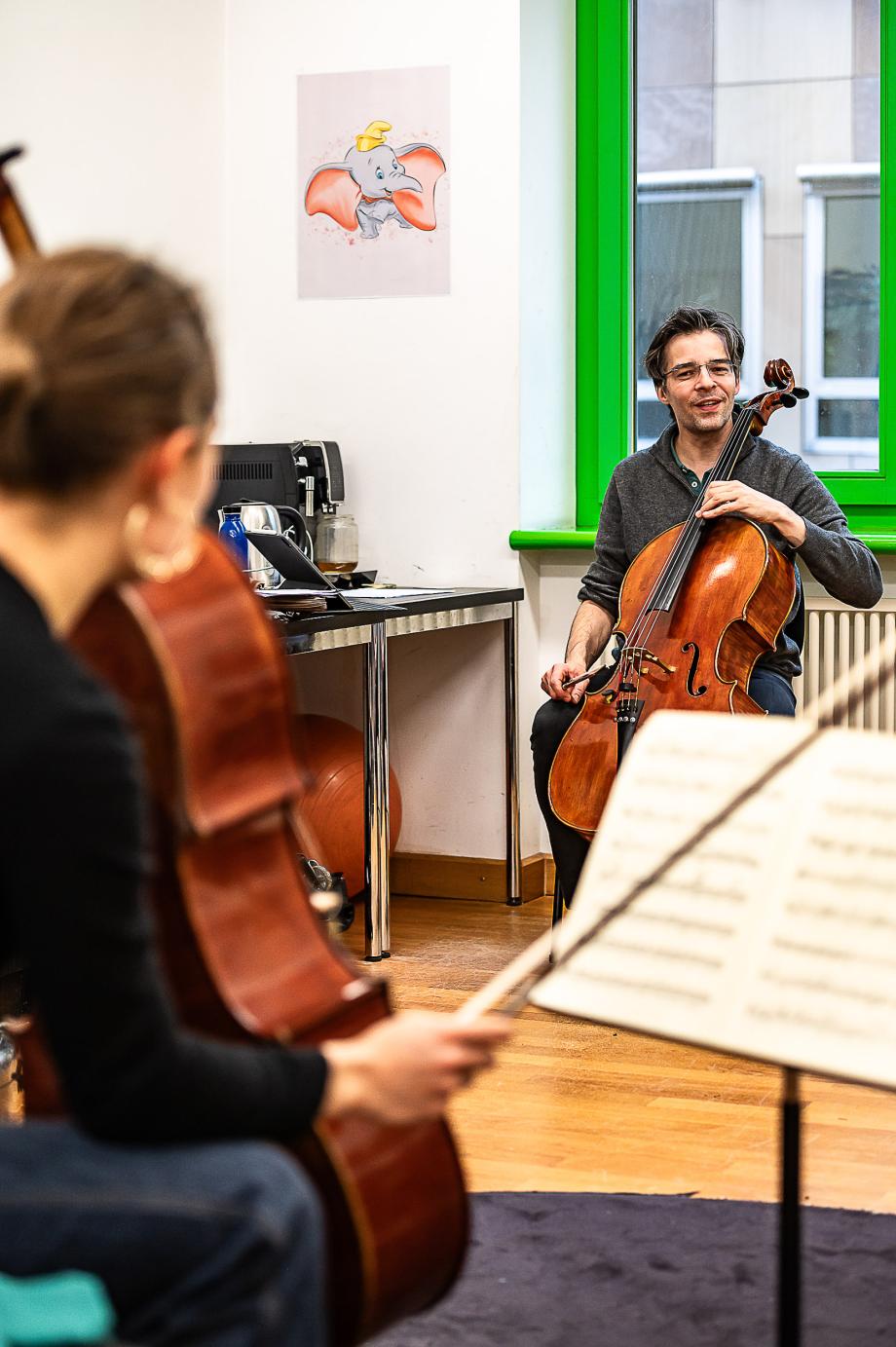 Vioncelloprofessor beim Einzelunterricht mit einer Studierenden. Die beiden sitzen sich gegenüber und befinden sich im Dialog. Im Hintergrund bildet der grelle Grünton des Fensterrahmens heraus.
