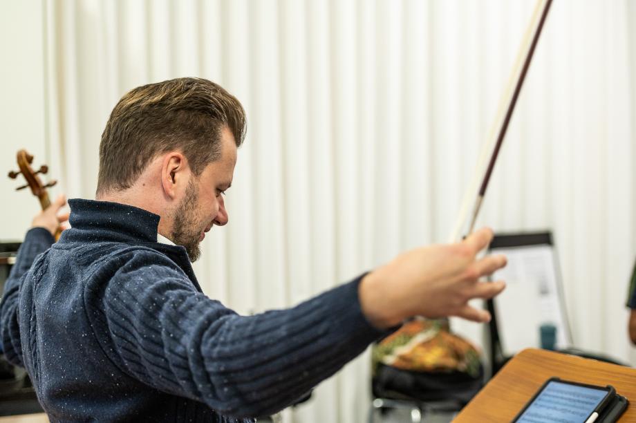 Violineprofessor Martin Fundamit einer auslandenen Geste. Beide Arme sind weit geöffnet. In der rechten Hand hält er die Violine.