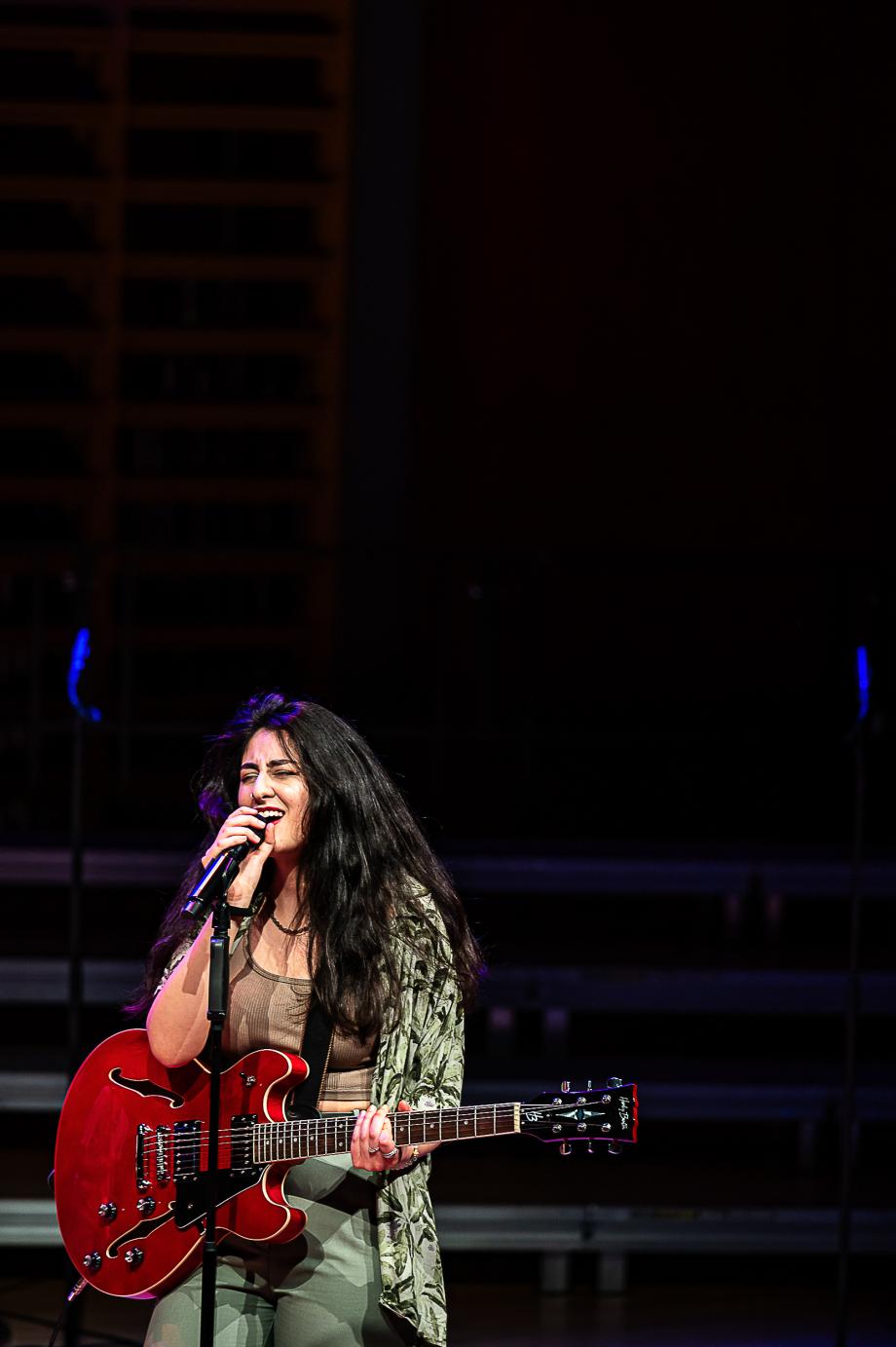 Singende Studierende mit roter E-Gitarre auf dem Konzertsaalpodium. Sie fokussiert sich auf das Mikrophon, die Augen sind geschlossen.