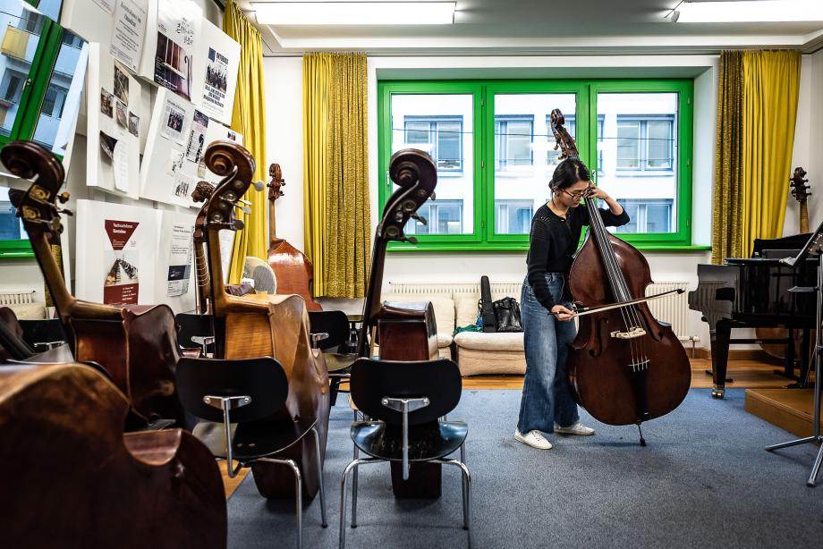 Musizierende Kontrabassstudentin im Unterrichtsraum. Neben ihr sind vier Kontrabässe auf die Stuhlfächen gelehnt.
