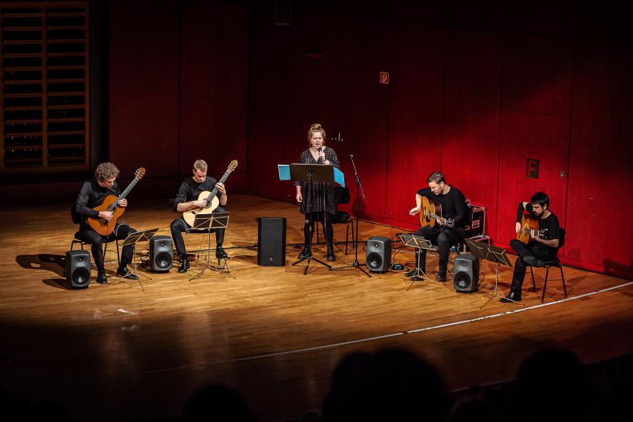 Gitarrenquartett mit einer Sängerin auf dem Konzertsaalpodium. Die Leitung hat Prof. Tillmann Reinbeck.