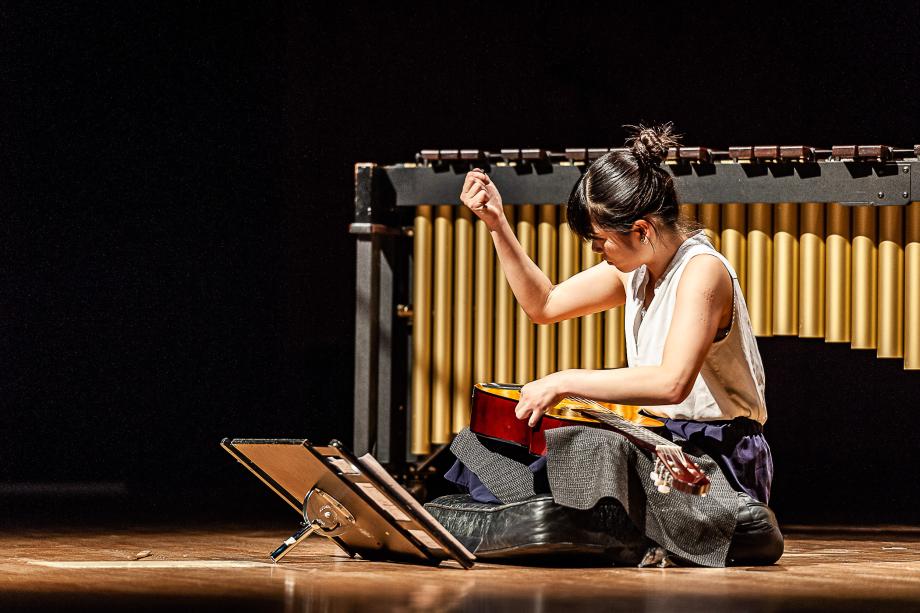 Schlagzeugstudnin sitzt im Schneidersitz auf dem Podium. Auf ihren Beinen liegt eine Gitarre, die rechte Hand nach oben zu einer Faust erhoben. Im Hintergrund ist ein Marimbaphon zu sehen, vor ihr ein kleiner Notenständer.