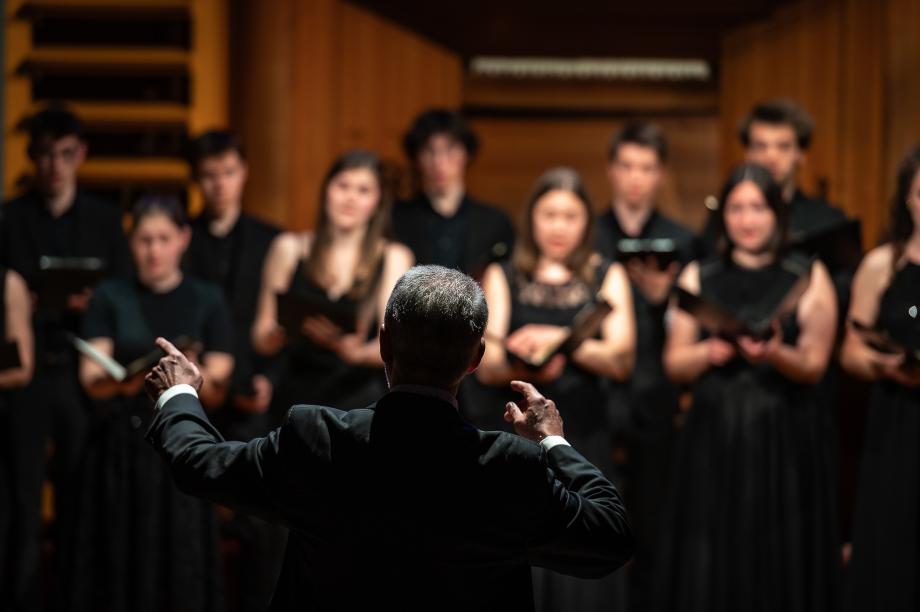 Prof. Denis Rouger dirigiert den Kammerchor. Der Fokus liegt auf seiner Person. Die Mitglieder sind nur teilweise zu erkennen.