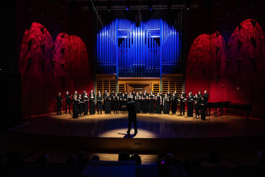Der Kammerchor unter der Leitung von Prof. Denis Rouger im Konzertsaal. Die Seitenwände und das Orgelprospekt sind atmosphärisch beleuchtet.
