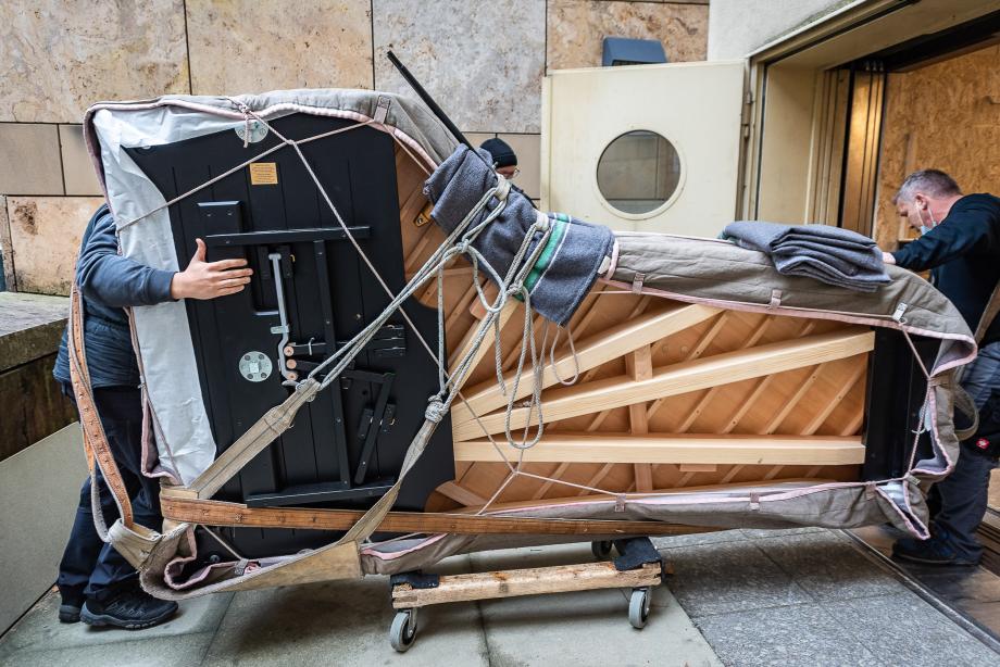 Anlieferung des neuen Konzertflügels vor dem Lastenaufzug der Hochschule. Der Flügel ist in unterschiedliche Filzdecken eingepackt und wird von zwei Monteuren auf einem Holzwagen transportiert.