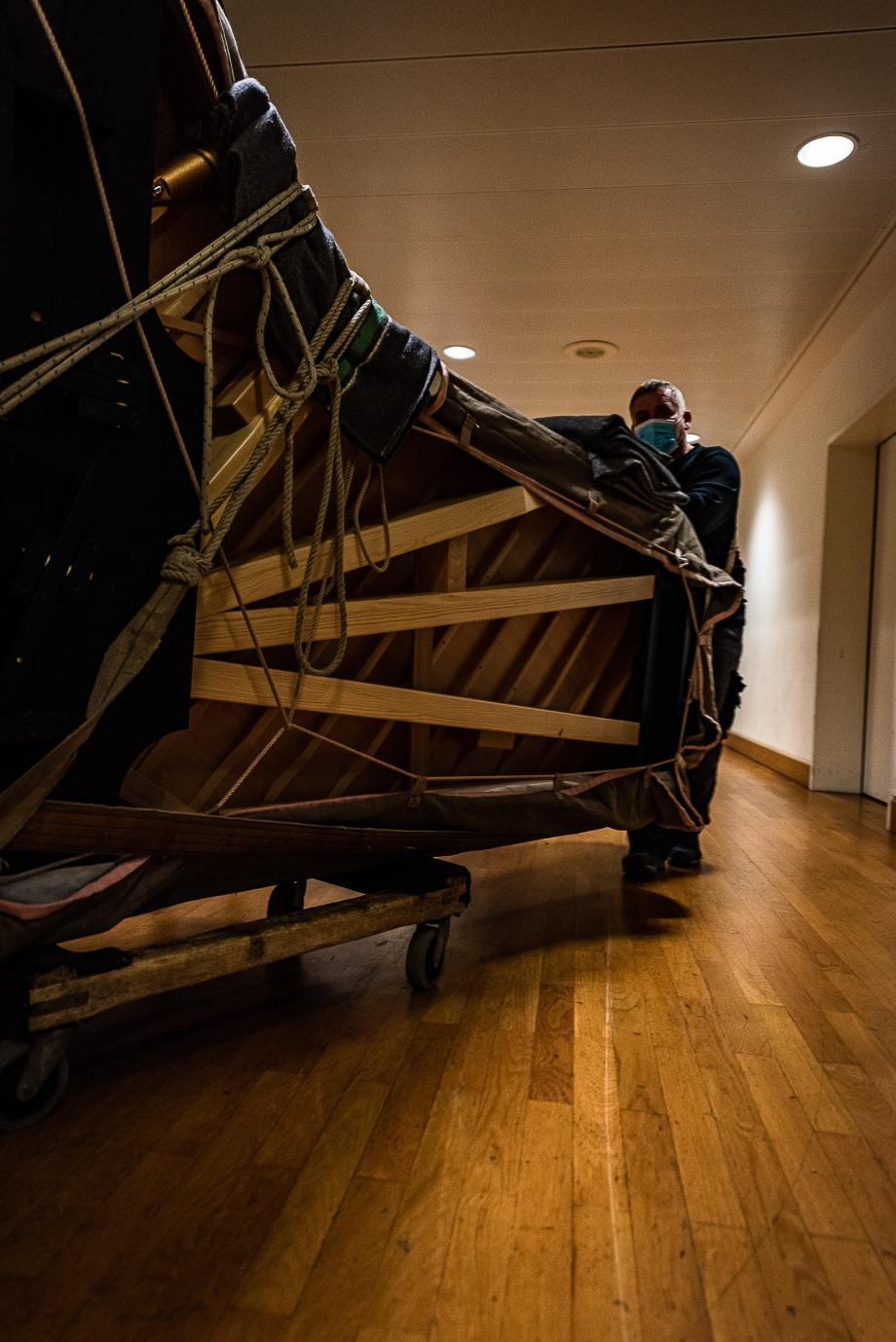 Anlieferungssituation des neuen Steinway-Konzertflügels. Das Instrumente ist hochkantig auf einem Holzwagen positioniert, mit Decken umgeben und von starken Seilen verspannt.