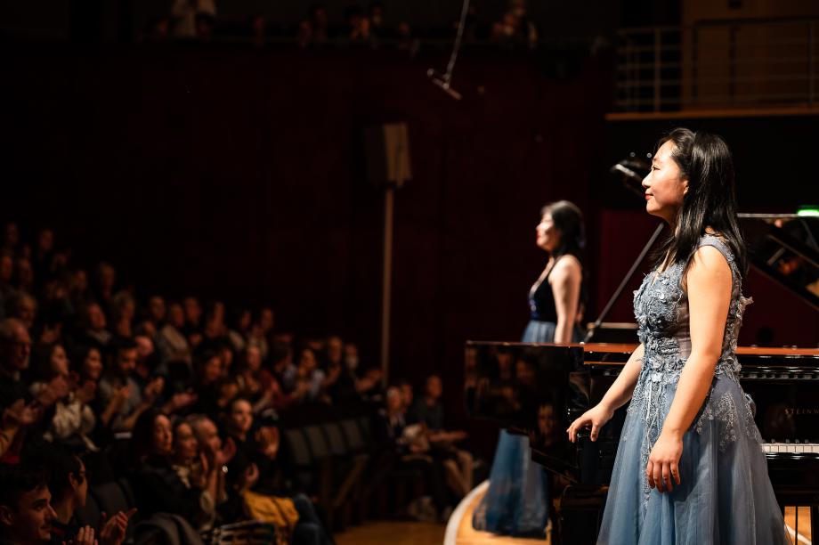 Seitliche Aufnahme eines Klavierduos bei ihrem Schlussapplaus. Die beiden Studierenden tragen blaue Abendkleider.