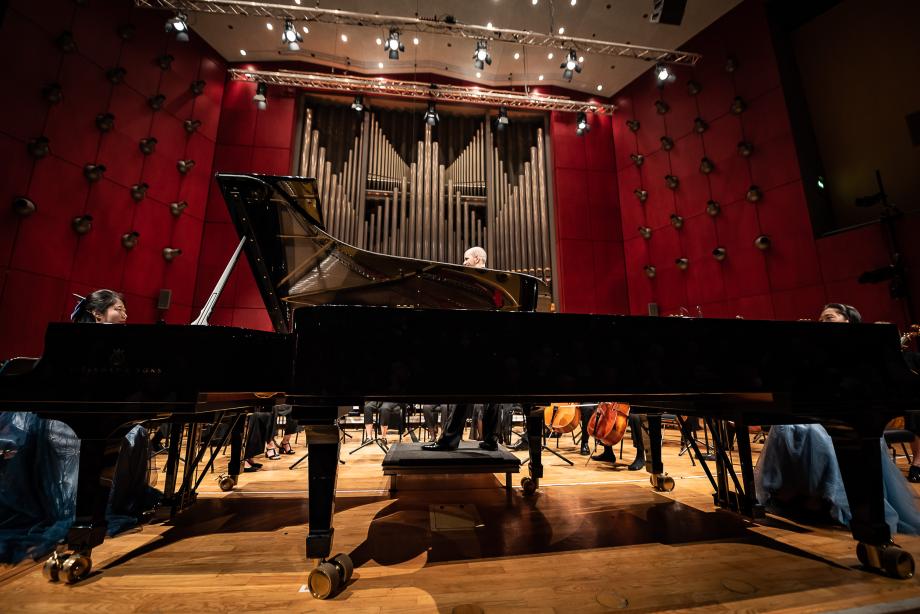 Detailaufnahme der beiden Konzertflügel und den Klaviersolistinnen in unmittelbarer Nähe der Bühnenkante. Im Hintergrund ist Prof. Rasmus Baumann und das Hochschulsinfonieorchester zu sehen.