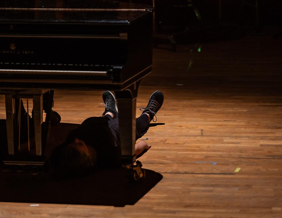 Im Rahmen einer Performnce liegt eine Studentin ausgestreckt unter dem Flügel auf dem Podiumsboden. Durch den Schatten des Flügels ist die Person kaum zu erkennen.