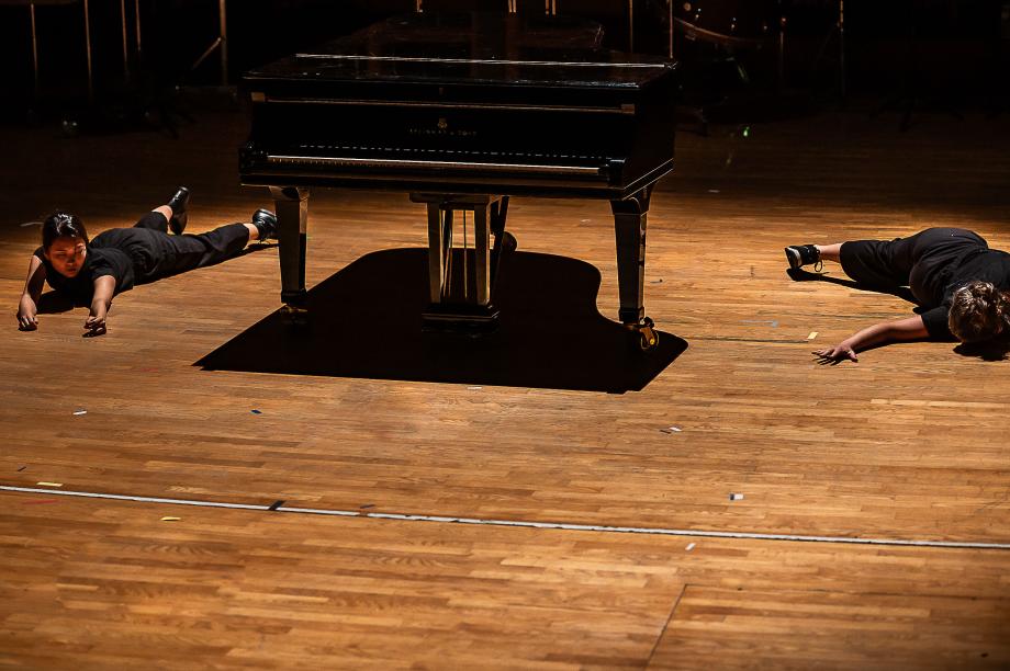 Zwei Studierende liegen links und recht neben dem Konzertflügel und drehen sich in verschiedenen Bewegungen. Der Flügel steht zentral und wirft einen klar umrandeten Flügelschatten auf das Podium.