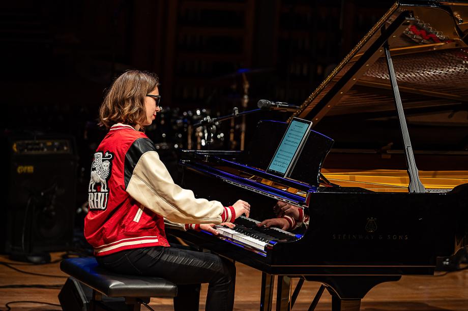 Portraitaufnahme einer Studetin am Flügel, mit farblich auffallender Lederjacke, schwarzer Hornbrille und einem Tablet.