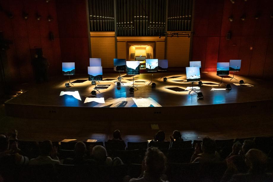 Blick aus dem Zuschaurraum auf die Installation von Nikolaus Koliusius. Es sind viele Fotografien von Anden zu sehen, die auf Notenpulten installiert und von jeweils einem Scheinwerfer illuminiert werden.