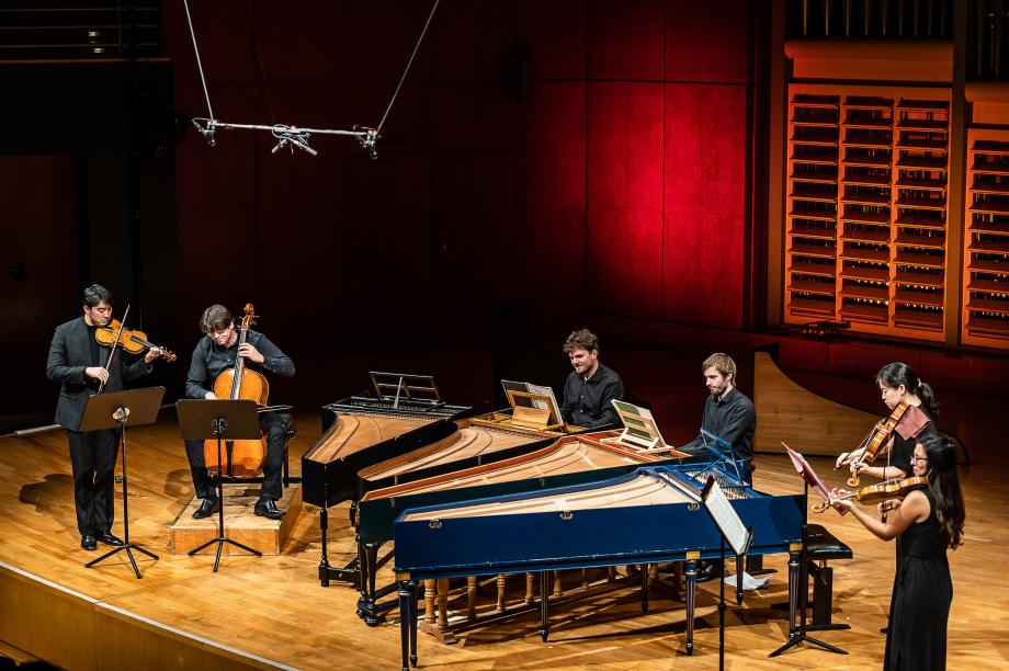 In der Bühnenmitte stehen vier Cembali aufgefächert. Zei Cembalisten musizieren, jeweils umgeben von Studierenden der Streicherklassen.