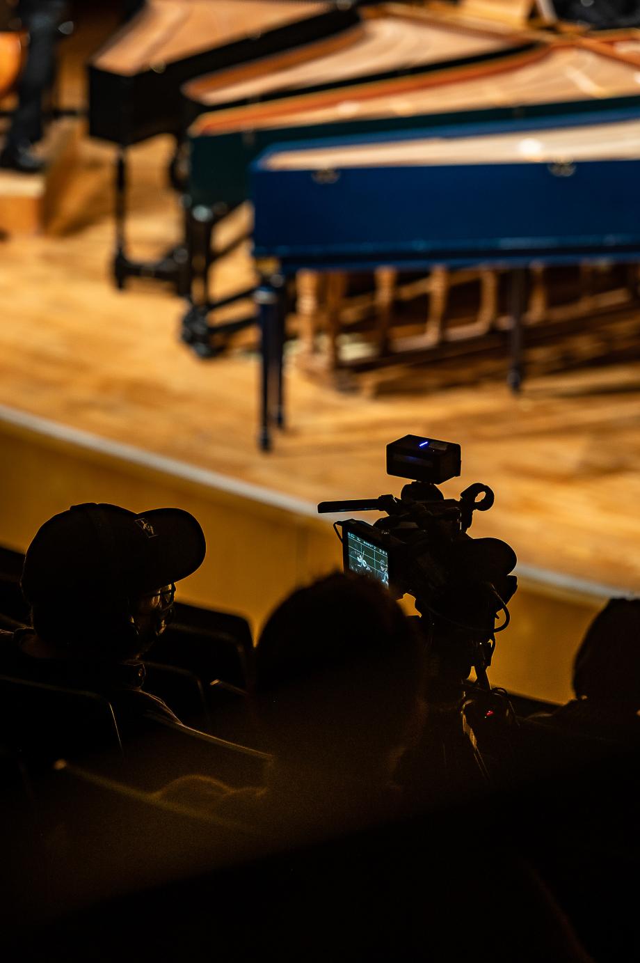 Blick aus dem Zuschauerraum auf die Bühne. Zu sehen ist ein Kameramann, der die Bühnenaktion verfolgt, am Bildrand stehen vier Cembali.