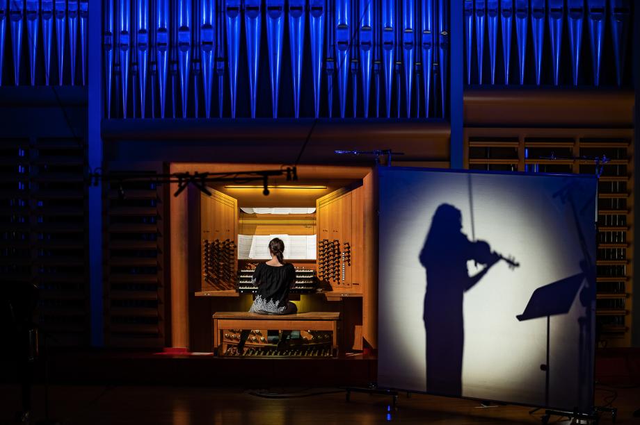 Blick auf den Orgeltisch im Konzertsaal. Daneben ist eine weiße Wand zu sehen und im Schatten eine geigende Studentin.
