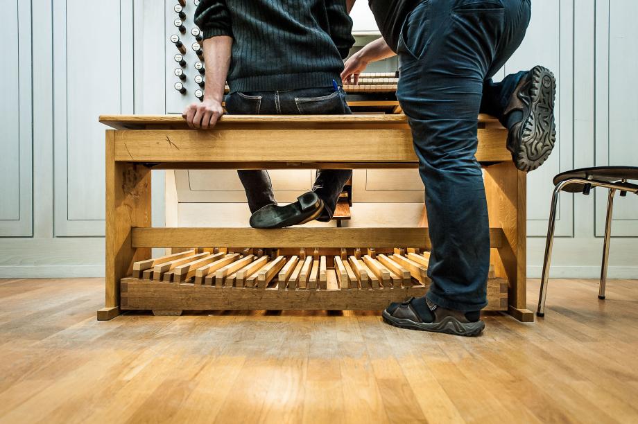 Detailaufnahme auf die Orgelbank und die Fußpedale einer Orgel. Zu sehen sind die Füße des Studierenden und die Lehrers, der ein Knie auf der Orgelbank aufgelegt hat.