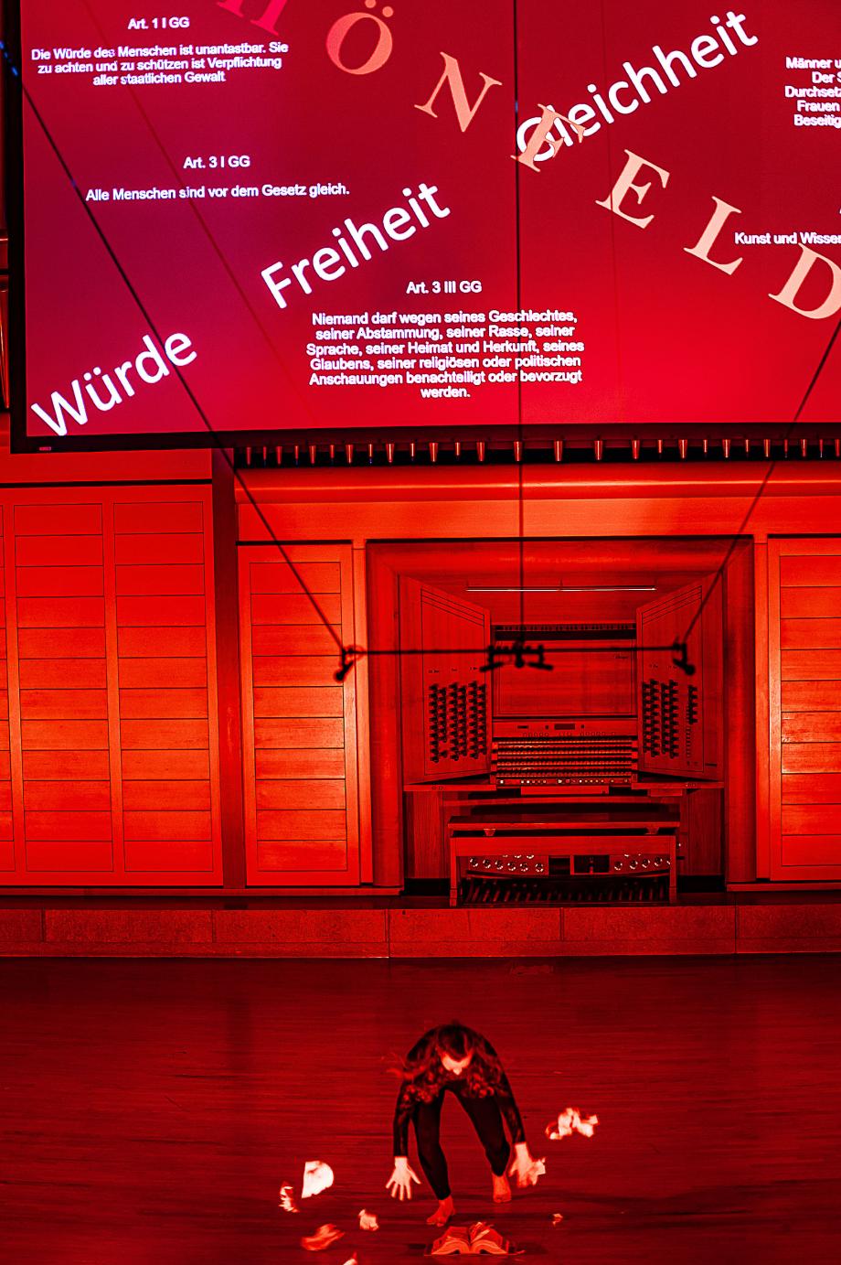 Der Konzertsaal ist in rot erleuchtet. Auf dem Podium befindet sich eine Studierende, die Bücher und Zetterl neben sich verteilt. Vor dem Orgelproespekt hängt eine Leinwand, auf der verschiedene Freiheitsrechte dargestellt werden.